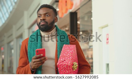 African american young man with Christmas present use phone walk in mall background Christmas tree feel happy smiling shopping internet face close up portrait mobile technology cell slow motion