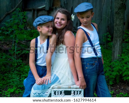 A little girl with two brothers sitting on a chair in a farm surrounded by greenery and a wooden barn