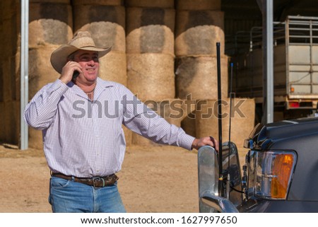 Smiling farmer on cell phone near truck with antennas. Hay bales stacked in shed in background