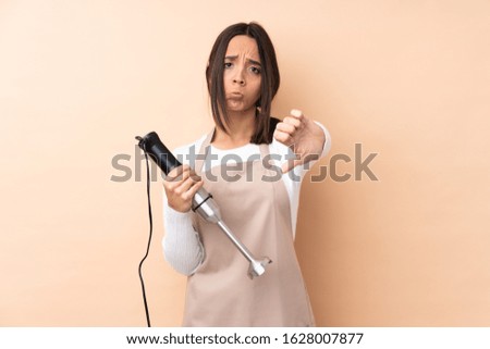 Young brunette girl using hand blender over isolated background showing thumb down with negative expression