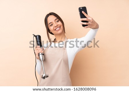 Young brunette girl using hand blender over isolated background making a selfie