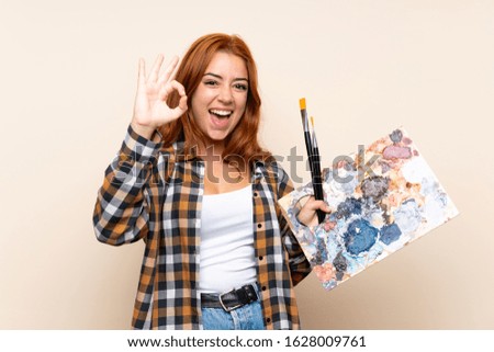 Teenager redhead girl holding a palette over isolated background showing an ok sign with fingers