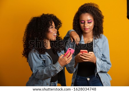 Image of emotional excited afro young women friends isolated over yellow wall background using mobile phones.