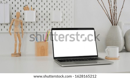 Cropped shot of modern designer workspace with mock up laptop, wooden figure, pencils and ceramic vase on white table with shelf on white wall background
