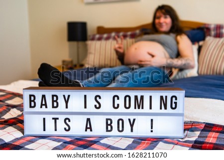 A selective focus view with a blurry woman in later stages of pregnancy lying in bed. and an LED light box displaying baby boy gender reveal message