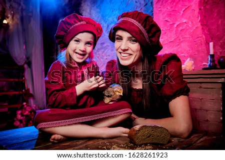 Easter and family. Mother and daughter in maroon or red dresses of village shepherdesses and small chicken in rural wooden interior in studio. Colour beautiful light background of two female relatives