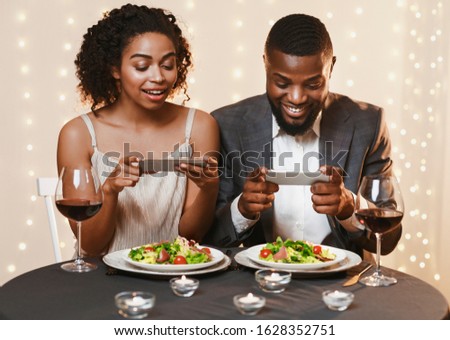 Young black couple sitting in restaurant and taking pictures of food with mobile phone, food bloggers celebrating Valentine's Day