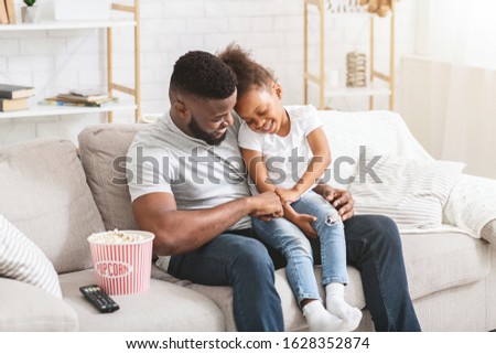 Cheery african dad tickling his little daughter while watching movie at home, copy space