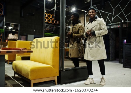 Handsome african american man posing  inside night club in black hat and beige coat.