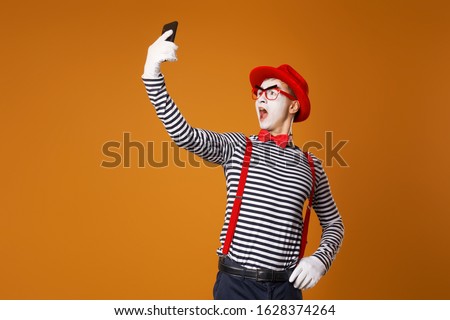 Young mime in white gloves and red hat takes selfie on orange background
