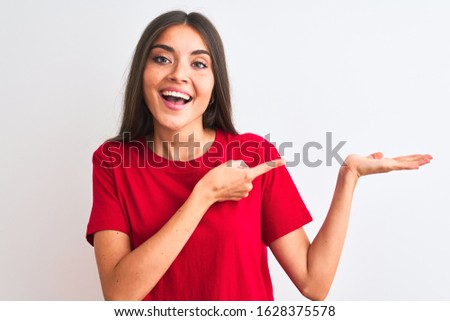 Young beautiful woman wearing red casual t-shirt standing over isolated white background amazed and smiling to the camera while presenting with hand and pointing with finger.