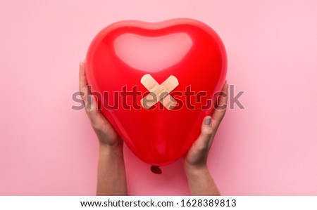 First Aid Band Plaster Strip Medical Patch glued on red heart balloon in woman hands on pink background. Heart broken, Love and Valentines day concept.