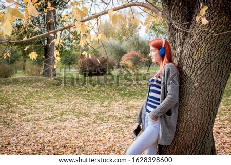 Young woman leaning on a tree while listening music over headphones and day dreaming in autumn park. 