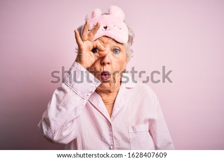 Senior beautiful woman wearing sleep mask and pajama over isolated pink background doing ok gesture shocked with surprised face, eye looking through fingers. Unbelieving expression.