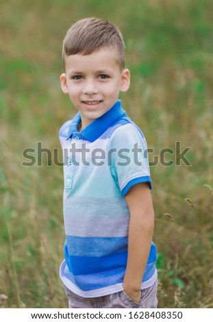Vertical color photography of cute little white boy posing for portrait standing outdoor in green summer landscape.