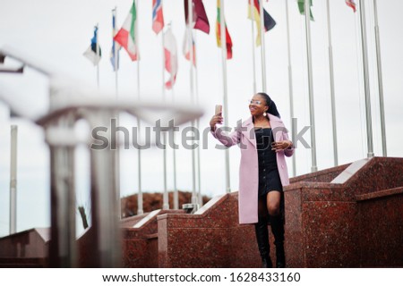 Young stylish beautiful african american woman in street, wearing fashion outfit coat, against flags of different countries of the world, with mobile phone at hand.