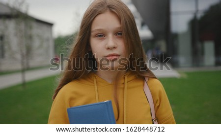 Joyful teenage student ready for school holding a book abd backpack staying in the school courtyard. Portrait of a pretty smart girl smiling outdoors near in the green area.