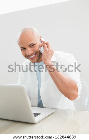 Portrait of a casual smiling young man using cellphone and laptop at desk against white background