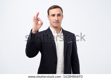 Portrait of cheerful italian young man in suit showing okay gesture, isolated over white background