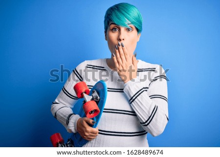 Young skater woman with blue fashion hair holding skateboard over blue isolated background cover mouth with hand shocked with shame for mistake, expression of fear, scared in silence, secret concept
