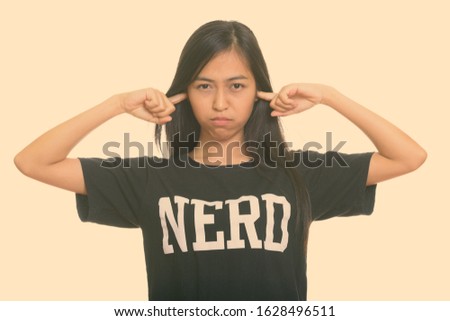 Studio shot of young Asian teenage nerd girl covering ears with fingers