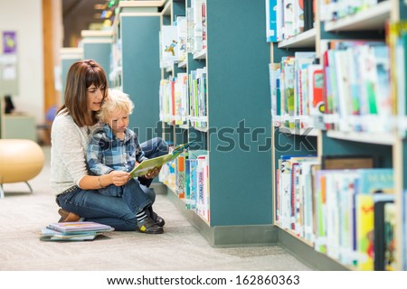 Full length of teacher and boy reading book by bookshelf in library