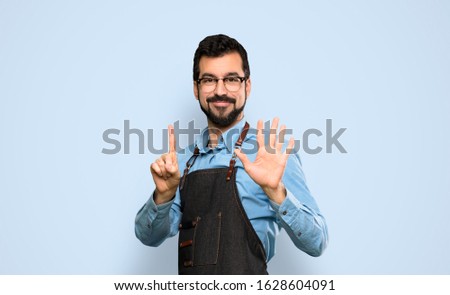 Man with apron counting six with fingers over isolated blue background