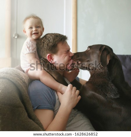 Family portrait of young handsome father with cute smiling little infant sitting on his shoulders and pet labrador retriever licking his face and hugging, side view