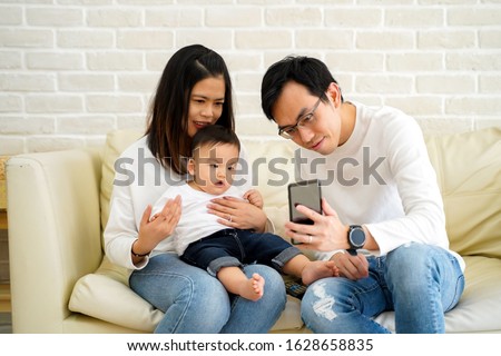 Cheerful young Asian family with son watching video on mobile phone and sitting on sofa couch at home, parents and kid enjoying cartoon movie
