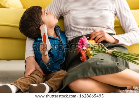 cropped view of woman sitting on floor with flowers near adorable son holding mothers day card with heart symbol