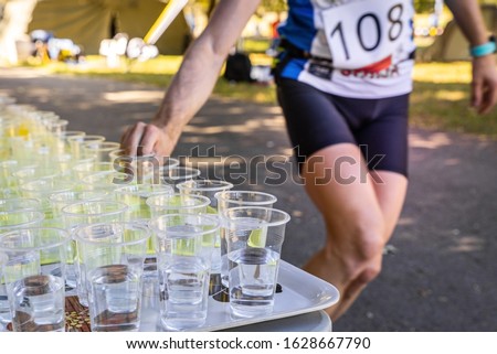 Runner with race number grabbing plastic cup with water at the refreshment station on the race. 
Focus on cups.  Royalty-Free Stock Photo #1628667790