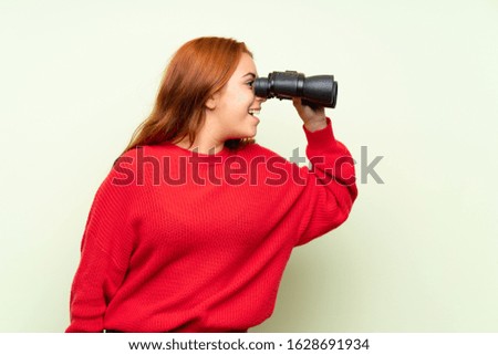 Teenager redhead girl with sweater over isolated green background with black binoculars