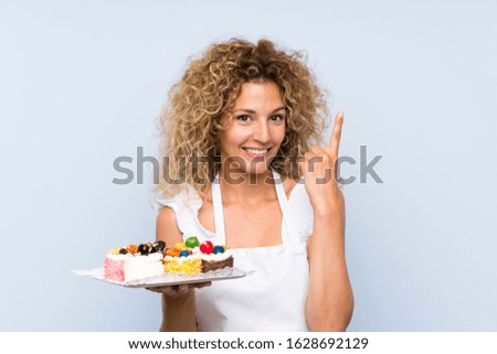 Young blonde woman with curly hair holding lots of different mini cakes pointing up a great idea