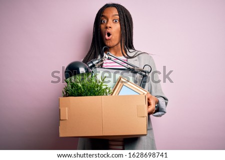 Young african american worker girl holding office cardboard fired from business job scared in shock with a surprise face, afraid and excited with fear expression