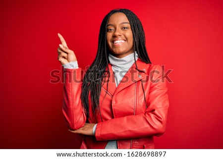 Young african american woman wearing cool fashion leather jacket over red isolated background with a big smile on face, pointing with hand and finger to the side looking at the camera.