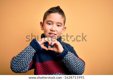 Young little boy kid wearing winter sweater over yellow isolated background smiling in love showing heart symbol and shape with hands. Romantic concept.