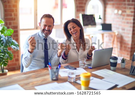 Middle age beautiful business workers working together using laptop at the office very happy and excited doing winner gesture with arms raised, smiling and screaming for success. Celebration concept.