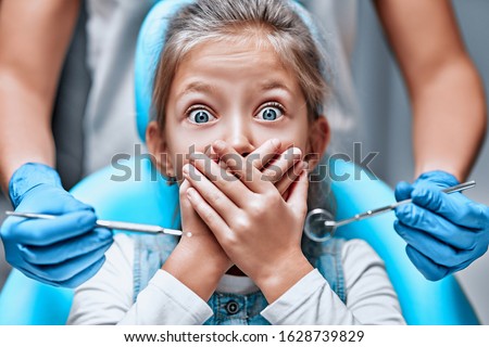 Close up view of a little girl looking scared and terrified screaming covering her mouth from the dentists with medical tools. Front view Royalty-Free Stock Photo #1628739829