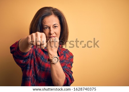 Middle age beautiful woman wearing casual shirt standing over isolated yellow background Punching fist to fight, aggressive and angry attack, threat and violence