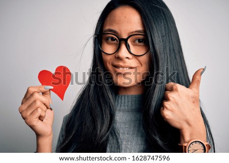 Young beautiful chinese woman holding paper heart over isolated white background happy with big smile doing ok sign, thumb up with fingers, excellent sign