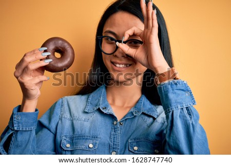 Young beautiful chinese woman holding chocolate donut over isolated yellow background with happy face smiling doing ok sign with hand on eye looking through fingers