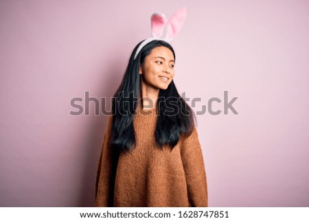 Young beautiful chinese woman wearing bunny ears standing over isolated pink background looking away to side with smile on face, natural expression. Laughing confident.