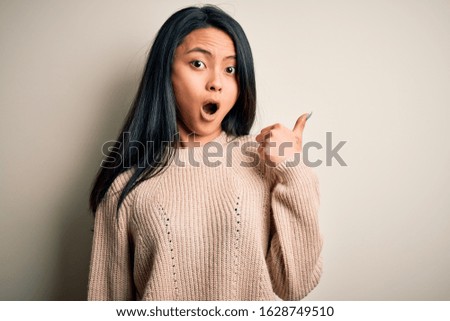 Young beautiful chinese woman wearing casual sweater over isolated white background Surprised pointing with hand finger to the side, open mouth amazed expression.