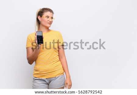 young pretty latin woman feeling sad, upset or angry and looking to the side with a negative attitude, frowning in disagreement with a smartphone against white wall