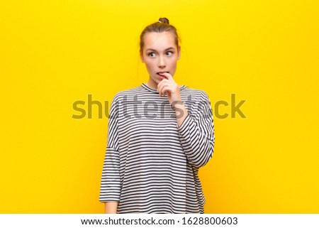young blonde pretty girl with surprised, nervous, worried or frightened look, looking to the side towards copy space against yellow wall