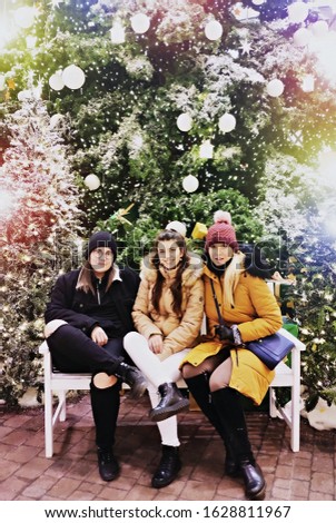 Three smiling sisters dressed cap and winter jacket sitting each other on Christmas tree on background. Merry Christmas, happy holidays, family and friendship concept.