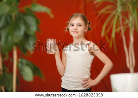cute child, pretty blonde little girl of 8 years old, drinks water from a glass on a red background surrounded by plants