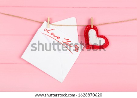 Paper envelope with fabric heart and text Love You hanging on pink wooden background