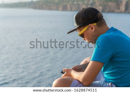 White kid using smartphone sitting outdoor isolated at blurry blue sea water background. Horizontal color photography.