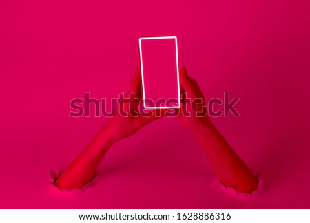 Female hands holding smartphone with neon glow frame through the torn holes. Pink light. Creative art